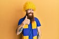 Redhead man with long beard football hooligan cheering game wearing funny wig doing happy thumbs up gesture with hand