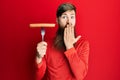 Redhead man with long beard holding fork with pork sausage covering mouth with hand, shocked and afraid for mistake Royalty Free Stock Photo