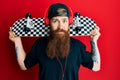 Redhead man with long beard holding skate wearing headphones relaxed with serious expression on face Royalty Free Stock Photo