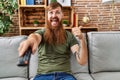 Redhead man with long beard holding television remote control pointing thumb up to the side smiling happy with open mouth Royalty Free Stock Photo