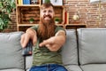 Redhead man with long beard holding television remote control smiling happy pointing with hand and finger Royalty Free Stock Photo