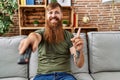 Redhead man with long beard holding television remote control smiling happy pointing with hand and finger to the side Royalty Free Stock Photo