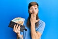 Redhead man with long beard holding wallet euro banknotes covering mouth with hand, shocked and afraid for mistake Royalty Free Stock Photo