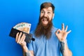 Redhead man with long beard holding wallet euro banknotes doing ok sign with fingers, smiling friendly gesturing excellent symbol Royalty Free Stock Photo