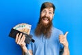 Redhead man with long beard holding wallet euro banknotes pointing thumb up to the side smiling happy with open mouth Royalty Free Stock Photo