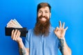 Redhead man with long beard holding wallet united kingdom pounds doing ok sign with fingers, smiling friendly gesturing excellent