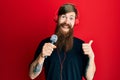 Redhead man with long beard singing song using microphone and wearing headphones pointing thumb up to the side smiling happy with Royalty Free Stock Photo