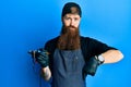 Redhead man with long beard tattoo artist wearing professional uniform and gloves pointing down looking sad and upset, indicating