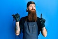 Redhead man with long beard tattoo artist wearing professional uniform and gloves pointing up looking sad and upset, indicating