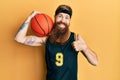 Redhead man with long beard wearing basketball uniform holding ball smiling happy and positive, thumb up doing excellent and Royalty Free Stock Photo