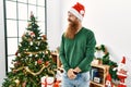 Redhead man with long beard wearing christmas hat by christmas tree looking away to side with smile on face, natural expression Royalty Free Stock Photo