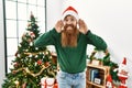 Redhead man with long beard wearing christmas hat by christmas tree trying to hear both hands on ear gesture, curious for gossip