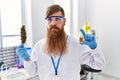 Redhead man with long beard working at scientist laboratory holding weed and cbd oil relaxed with serious expression on face
