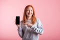 Redhead teenager girl points with finger at smartphone blank screen in studio on pink background. Royalty Free Stock Photo