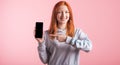 Redhead teenager girl points with finger at smartphone blank screen in studio on pink background. Royalty Free Stock Photo