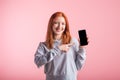 Redhead teenager girl points with finger at smartphone blank screen in studio on pink background. Royalty Free Stock Photo