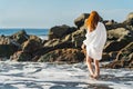 Redhead tender girl stand barefoot in the surf white foam on the beach near rocks. Woman in a white dress on the coast