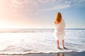 Redhead tender girl stand barefoot in the surf white foam on the beach. Woman in a white dress on the coast of an ocean