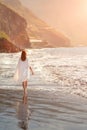 Redhead tender girl walks barefoot in the surf white foam on the beach. Woman in a white dress on the coast of an ocean