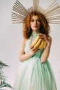 Redhead woman with accessory on head posing with bananas on grey