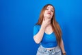 Redhead woman standing over blue background touching painful neck, sore throat for flu, clod and infection