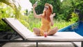 Redhead woman in yellow dress lounges by blue pool, tropical plants surround. Sips refreshing watermelon drink, snaps