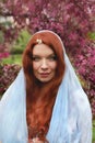 redhead young woman with green eyes and freckles wearing blue cape with cherry flowerm on her forehead in rose cherry