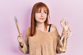 Redhead young woman holding small wooden manikin and painter brushes smiling looking to the side and staring away thinking Royalty Free Stock Photo