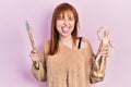 Redhead young woman holding small wooden manikin and painter brushes sticking tongue out happy with funny expression