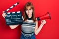 Redhead young woman holding video film clapboard and megaphone making fish face with mouth and squinting eyes, crazy and comical Royalty Free Stock Photo