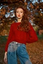 Redhead young woman in a red sweater walks in the park. Autumn beauty portrait of a fashionable Red-haired woman at sunset