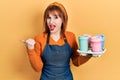 Redhead young woman wearing waitress apron holding take away cups of coffee pointing thumb up to the side smiling happy with open Royalty Free Stock Photo