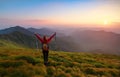 Redheaded girl athlete with a backpack and sticks stands on the green hillocks and looks at high mountain landscapes.