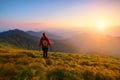 Redheaded girl athlete with a backpack and sticks stands on the green hillocks and looks at high mountain landscapes.