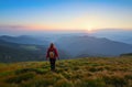 Redheaded girl athlete with a backpack and sticks stands on the green hillocks and looks at high mountain landscapes.