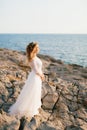 A refined graceful bride with a diadem stands on a rocky seashore, the sun's rays fall on her face