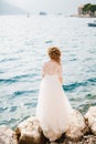 A refined graceful bride stands on a rocky seashore, in front of her the old town and boats, back view