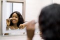 Reflection of pretty smiling African girl in mirror applying cosmetic cream on her face. Female putting moisturizer on