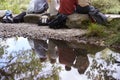 Reflection in stream of five young adult friends taking a break sitting on rocks during a hike, back view, low section