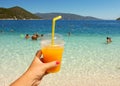 Refreshing in very hot summer day with fresh orange juice in women hand on the background of Antisamos beach, Sami