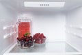 In the refrigerator on the shelf is a jug of juice, next to raspberries and cherries in transparent bowls