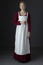 A Regency maid servant wearing a red linen dress with an apron and cap against a studio backdrop