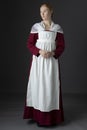 A Regency maid servant wearing a red linen dress with an apron and cap against a studio backdrop