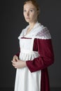 A Regency maid servant wearing a red linen dress with an apron and a lace modesty shawl against a studio backdrop