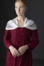 A Regency maid servant wearing a red linen dress with an apron and a lace modesty shawl against a studio backdrop