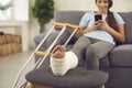 Woman with broken leg in plaster cast sitting on couch at home with mobile in hands Royalty Free Stock Photo