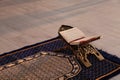 Rehal with open Quran and Muslim prayer beads on rug indoors.