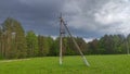 A reinforced concrete pole of a power line with electric wires stands in a meadow near the forest. A thunderstorm is coming from b