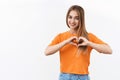 Relationship, emotions and youth concept. Portrait of cheerful, attractive blonde girl in orange t-shirt, show heart