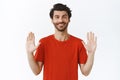 Relax, everything under control. Confident good-looking bearded guy in red t-shirt, raising hands up and smiling, asking